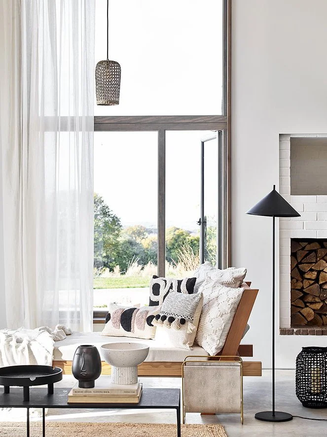 Modern living room with large windows, a wooden daybed with patterned pillows, a floor lamp, and a fireplace filled with stacked firewood.