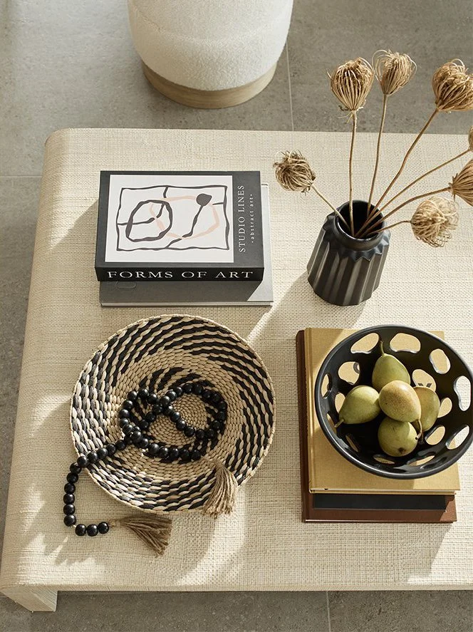 A beige table with a black and white book titled 'Forms of Art,' a black vase with dried beige flowers, a round woven basket with a black beaded necklace, and a black bowl filled with green pears, resting on two hardcover books.
