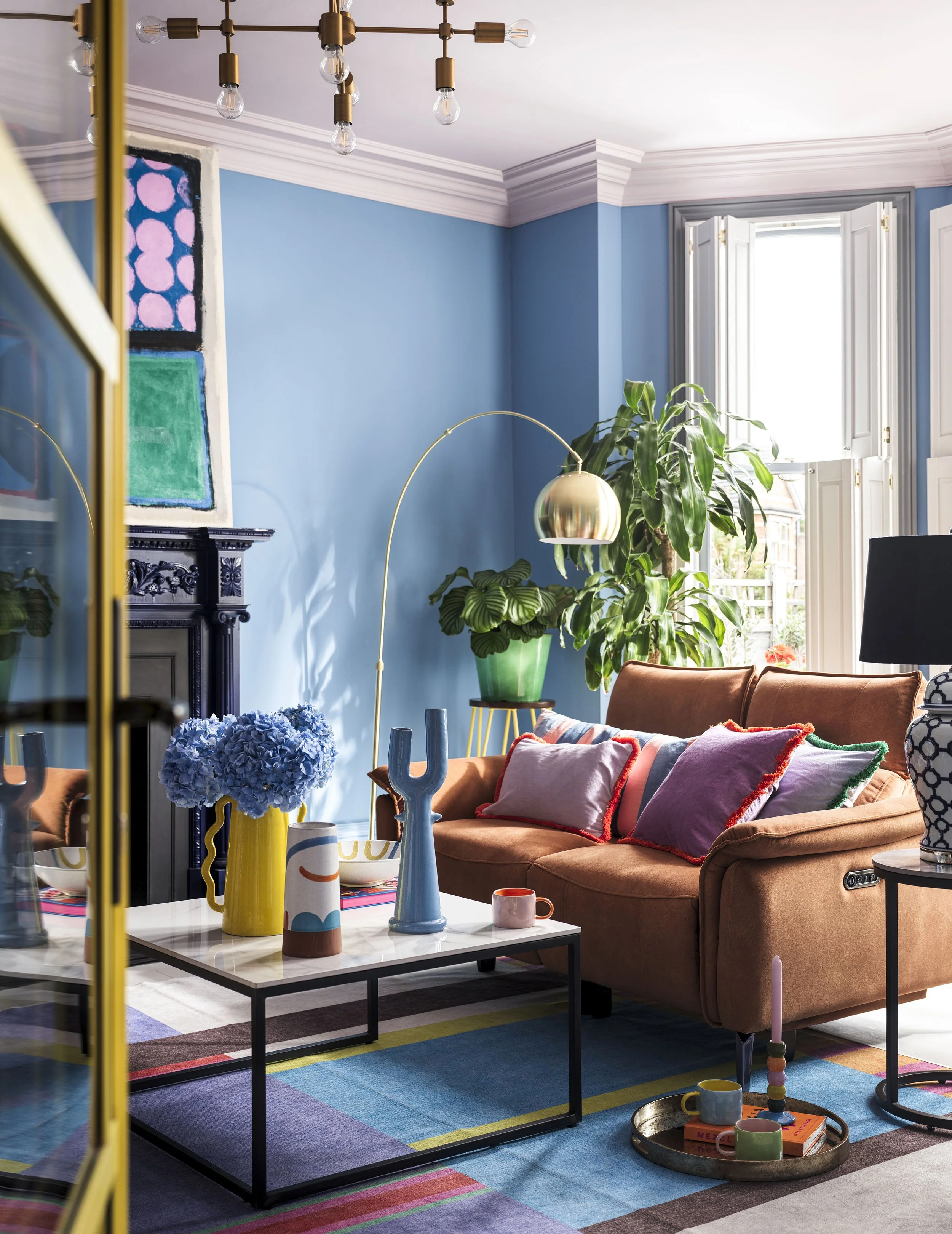 A colorful living room with a blue wall, brown velvet sofa with vibrant pillows, a white marble coffee table with decorative vases and cups, a multicolored rug, large green plants, and a window with white shutters. Gold and black accents are present 