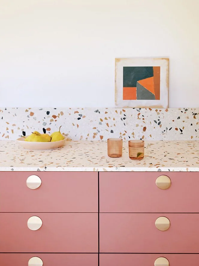 Pink kitchen cabinet with white knobs, terrazzo countertop with pears, pink glasses, and wall art on a white wall.