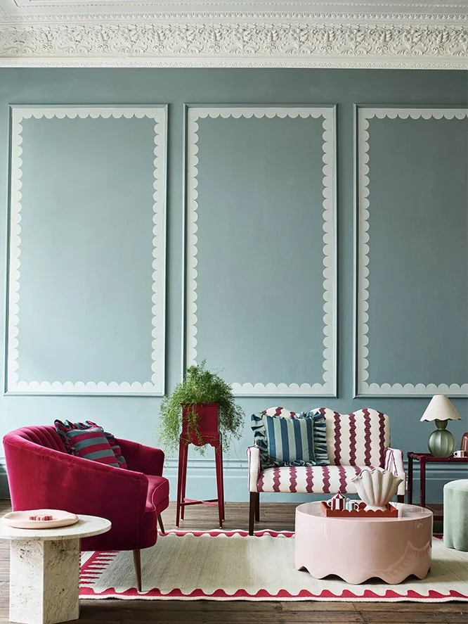 Interior living room with light blue walls, decorative picture frame moldings, and ornate crown molding. Features a red armchair with cushions, a white and red striped loveseat with striped pillows, a pink coffee table with a scalloped edge, a small 