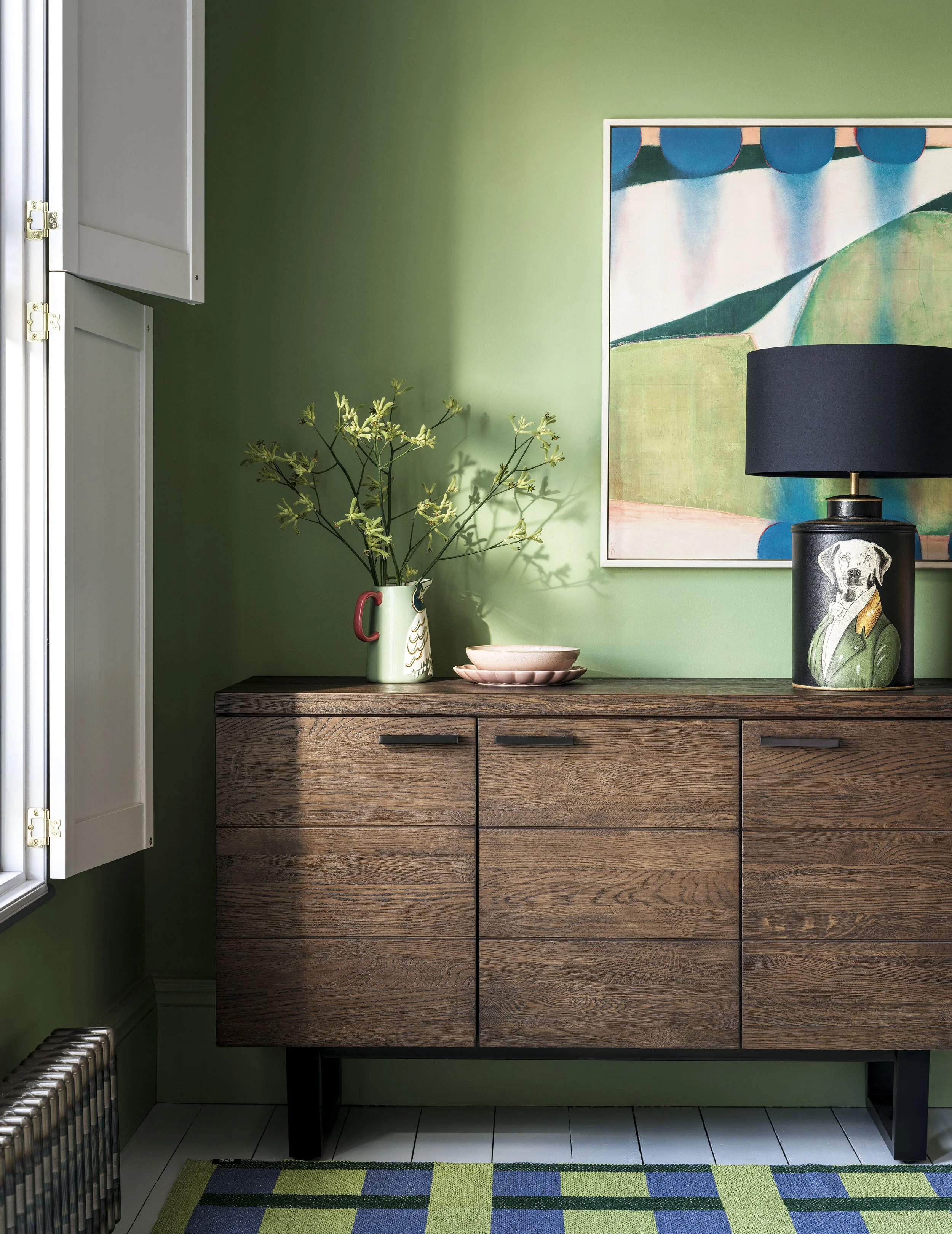 A wooden sideboard against a green wall, decorated with a potted plant, pink plates, and a lamp with a portrait of a dog wearing a green coat, with a colorful abstract painting hanging above.