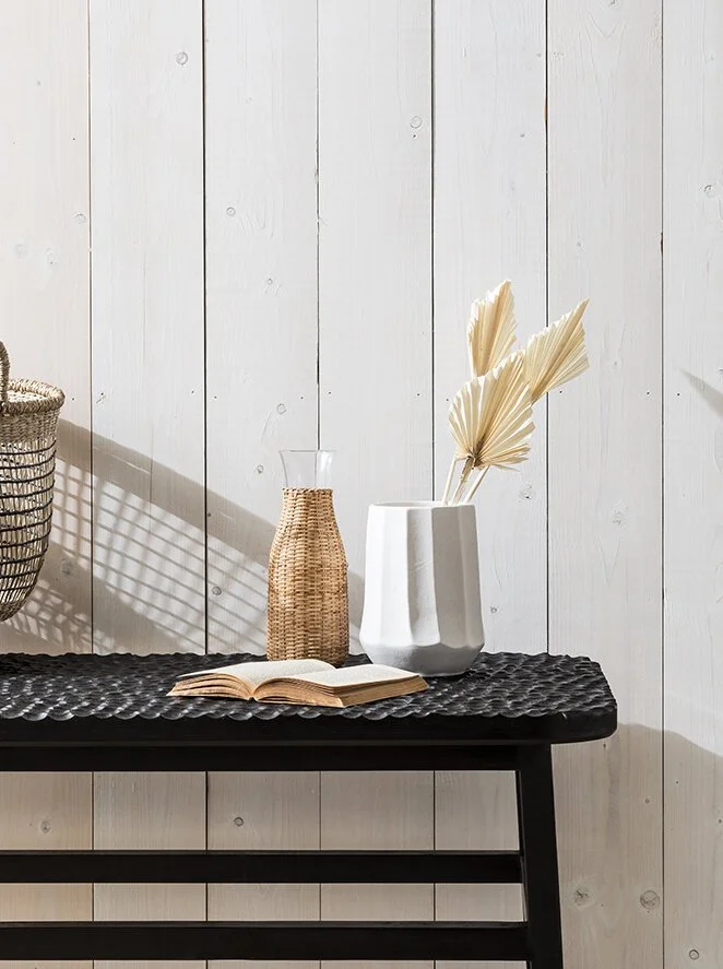 Decorative objects on a black textured table against a white wooden wall, including an open book, a beige woven vase, a white geometric vase with dried palm leaves, and a wicker basket to the side.