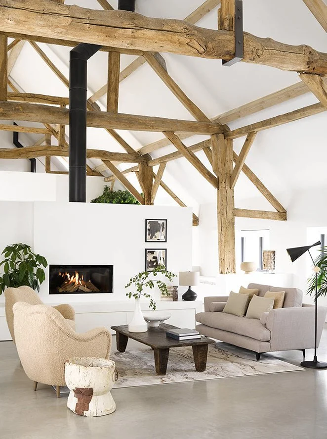 Modern living room with beige armchair and light gray sofa, rustic wooden beams on the ceiling, a fireplace, potted plants, and black and white artwork on the white wall.