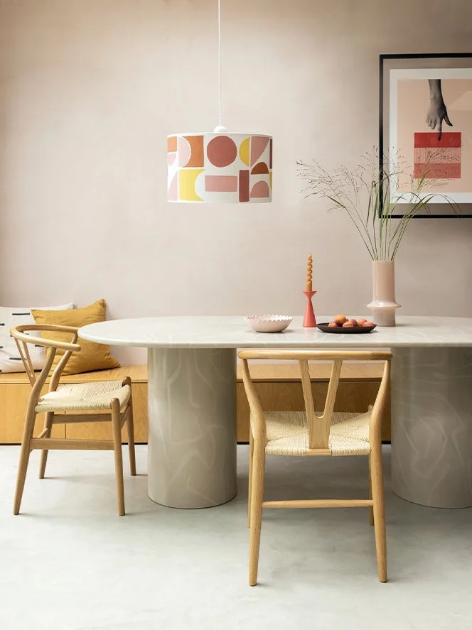 Modern dining room with a white marble table, wooden chairs with woven seats, a built-in bench with mustard cushions, a colorful geometric pendant lamp, a framed art piece, and a tall pink vase with branches.