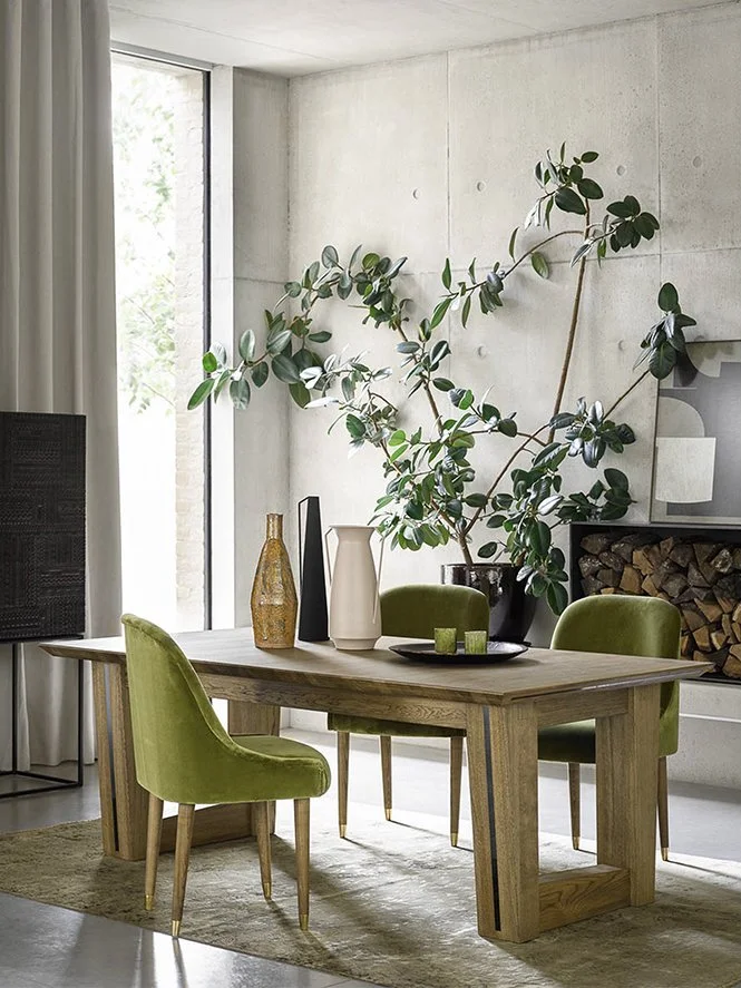 A dining room with a wooden table, green upholstered chairs, large houseplant, decorative vases, and a fireplace with firewood. Window with white curtains on the left.