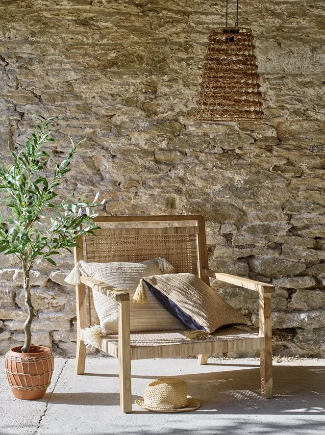 A wooden bench with cushions and pillows, a straw hat on the ground, a potted plant, a stone wall backdrop, and a woven hanging light fixture.