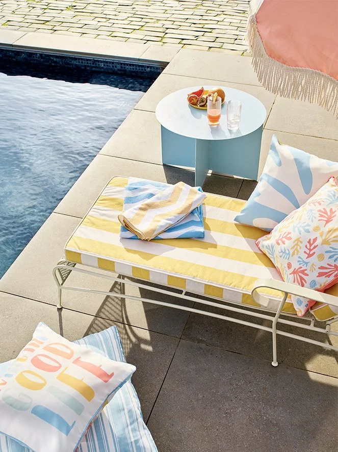Poolside outdoor space with a lounge chair, colorful pillows, yellow and white striped cushion, a folded towel, a round blue side table with drinks and snacks, and a pink patio umbrella with fringe near a swimming pool.