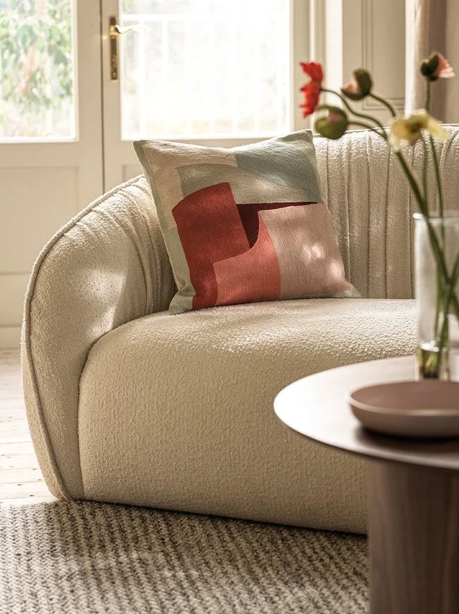 A cozy living room corner with a beige sofa, a colorful abstract pillow, a wooden side table with a bowl, and a glass vase of flowers, near a bright window.