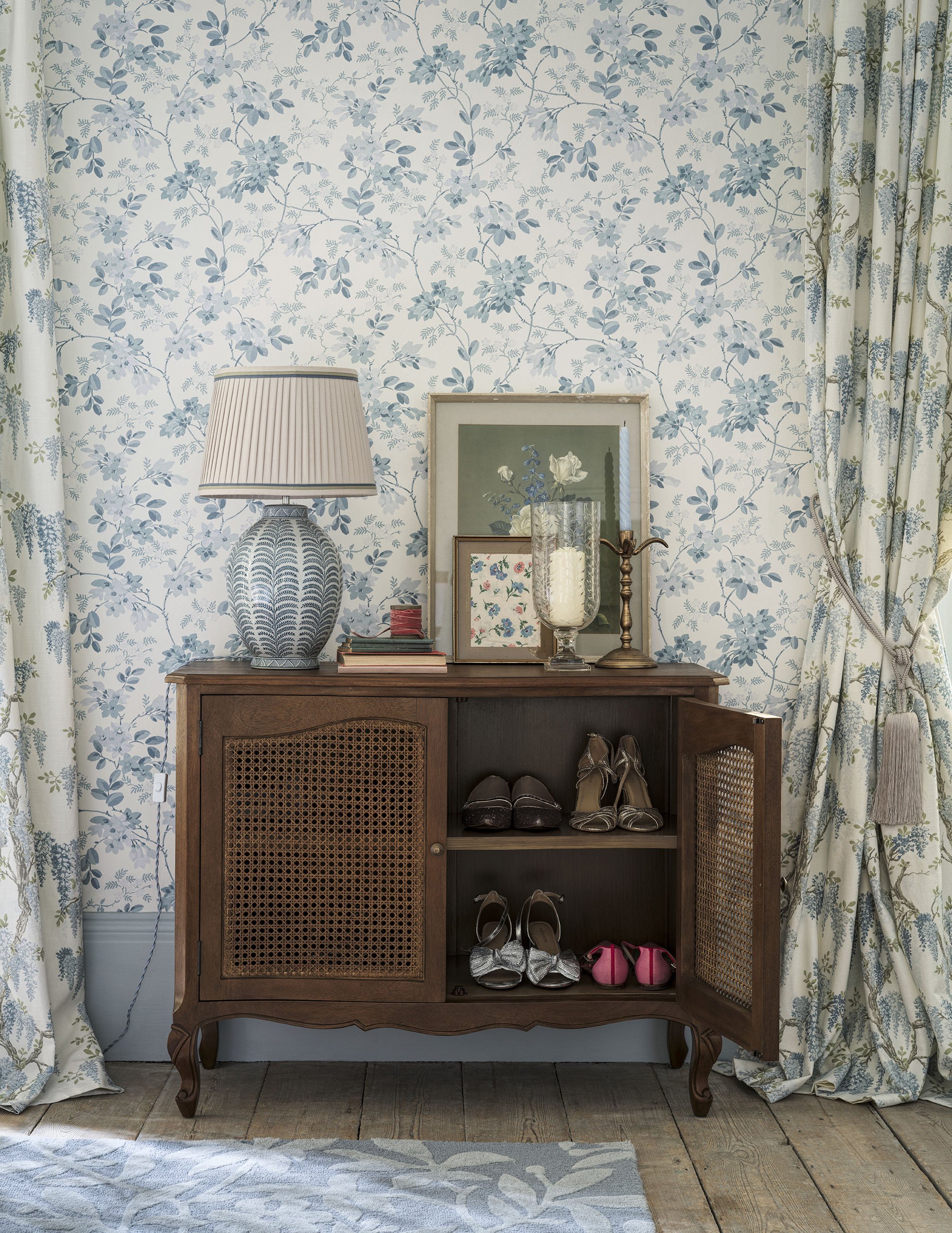 A wooden sideboard with open doors showing shoes inside, topped with a table lamp, framed photographs, a candle holder, and a large glass vase with a candle, in front of floral wallpaper and curtains.
