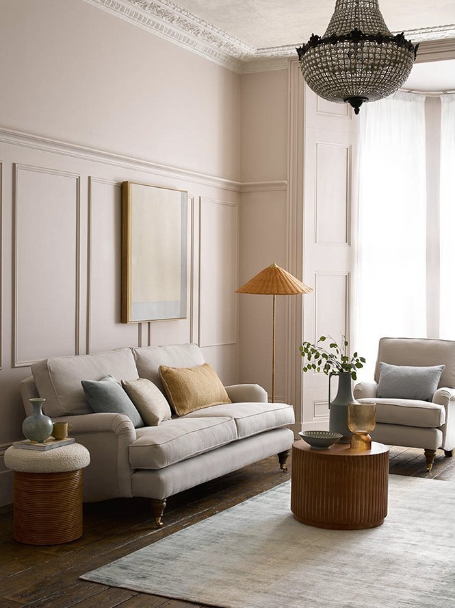 Living room with white sofa and armchair, beige pillows, a wooden round coffee table with vases and bowls, a floor lamp with a lampshade, a chandelier, and a window with sheer curtains.