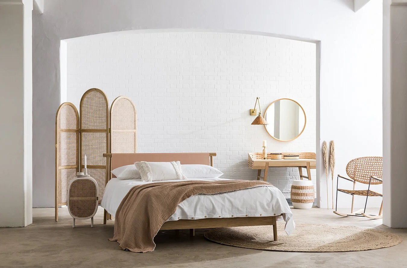 A minimalist bedroom with a white brick wall, beige and brown decor, a bed with white linens, a brown throw blanket, a wooden headboard, a rattan screen behind the bed, a round mirror above a wooden desk, a wicker basket, a rocking chair, and a beige