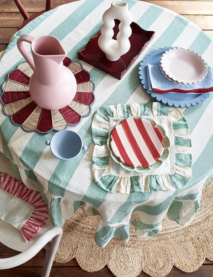 A round table with a green and white striped tablecloth, set with colorful plates and a pink pitcher on a woven placemat, with decorative vases and napkins.