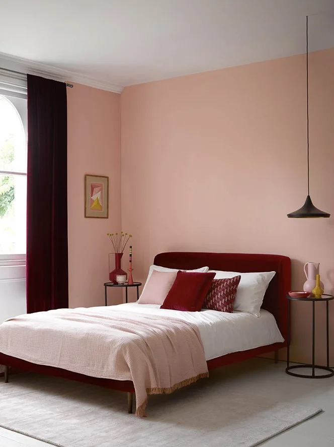 A bedroom with pink walls, a bed with white and pink bedding, and red pillows. There are two small black side tables with decorative vases, a framed artwork on the wall, a window with dark red curtains, and a black pendant light hanging from the ceil