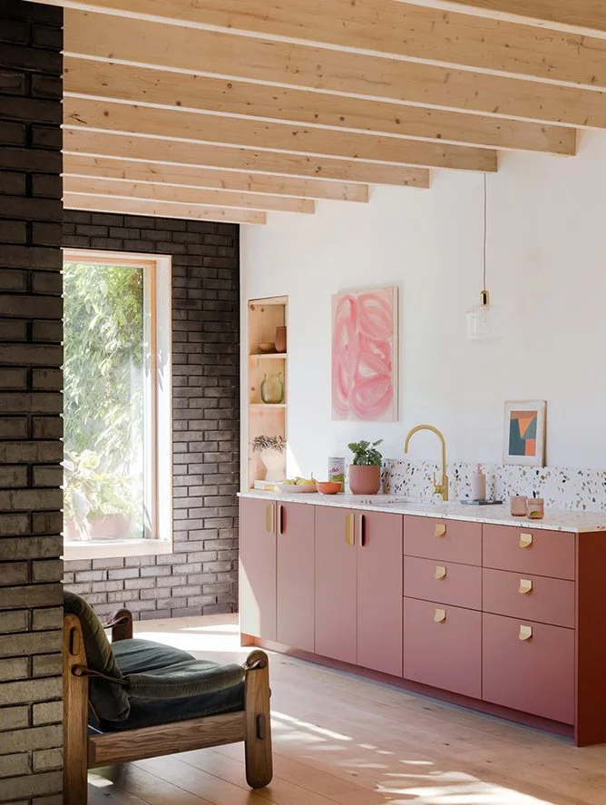 Modern kitchen with pink cabinetry, wooden ceiling beams, black brick wall, and natural light from a large window.
