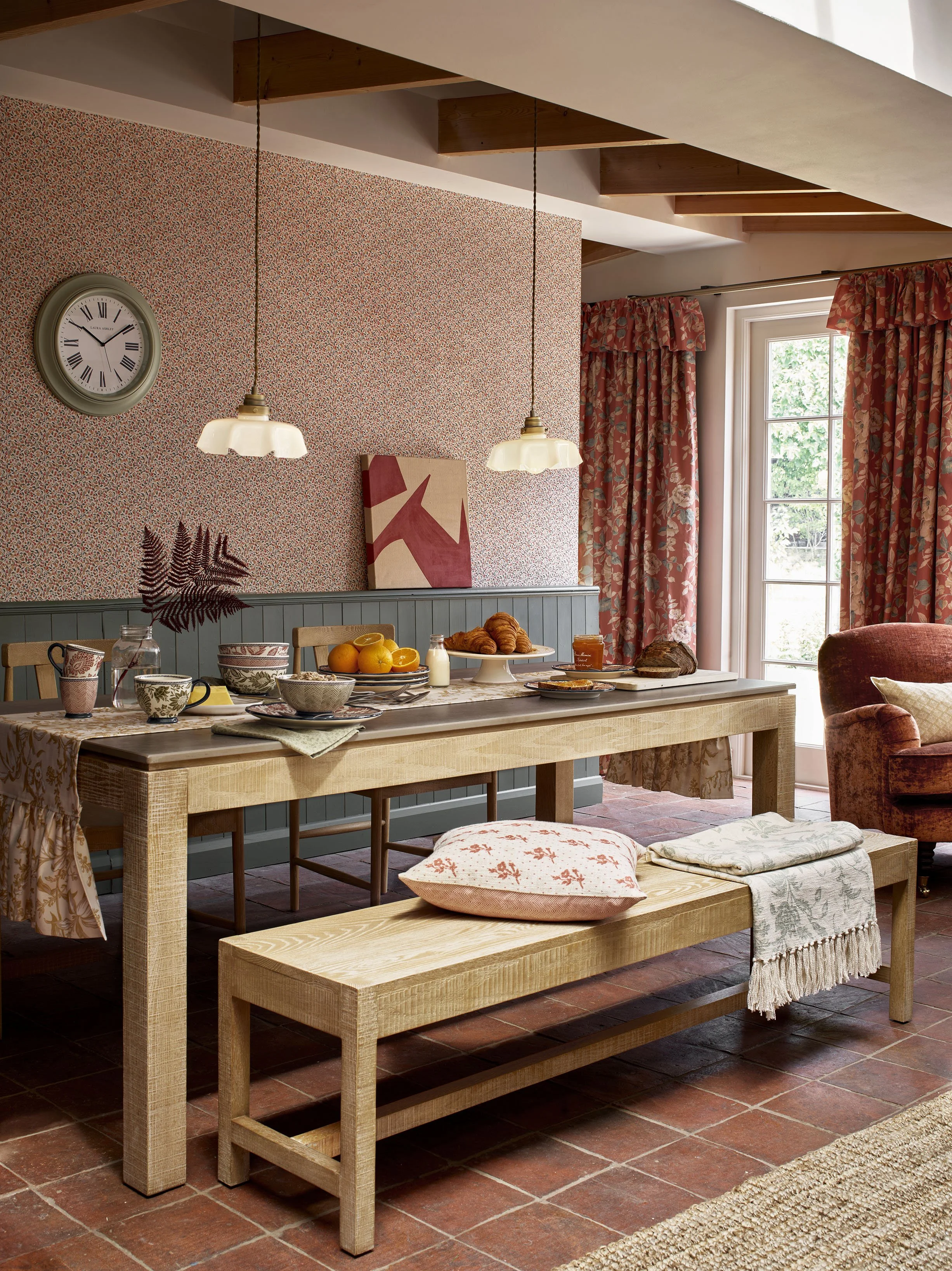 A cozy dining area with a wooden table set for breakfast, including bowls, cups, bread, croissants, oranges, and drinks. There is a bench with pillows underneath. The room features floral curtains, a patterned accent wall, a wall clock, and natural l