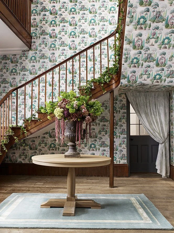 A foyer area with a staircase, wooden railing, floral wallpaper, a wooden table with a large floral arrangement, and a light blue rug. There's also a door with a drawn curtain.