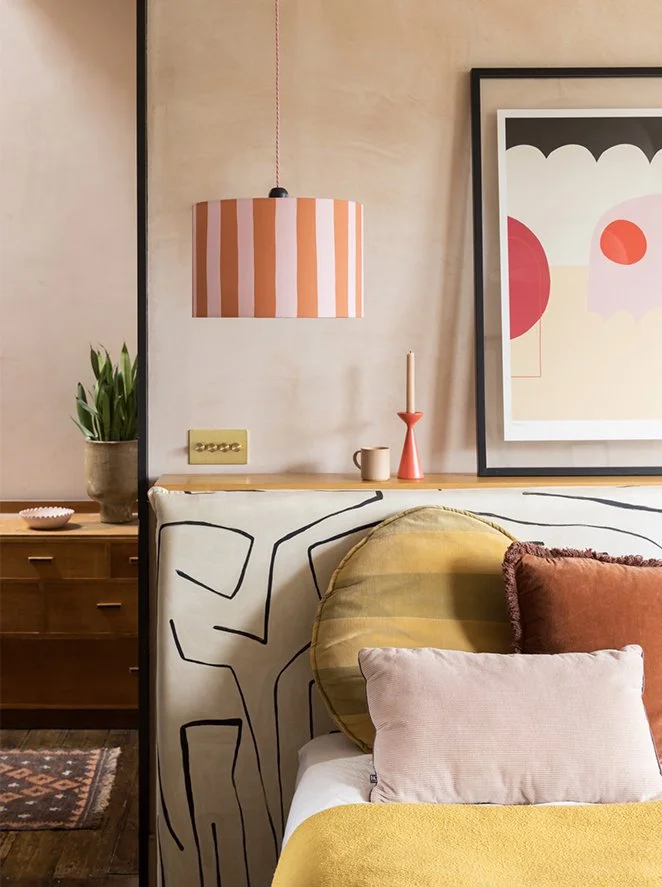 A cozy bedroom corner with a bed, colorful pillows, and modern decor. The wall features a large framed abstract art piece. A pendant light with a pink and white striped shade hangs from the ceiling. There is a wooden bedside table with a potted plant