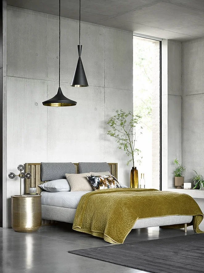 Modern bedroom with gray concrete walls, a large window, a bed with gray pillows and an olive-green blanket, a gold side table, pendant lights, and plants.