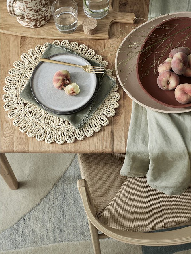 A wooden dining table set with a woven placemat, a stack of gray plates with a fork, and three peaches on top. There is a large pink bowl with more peaches and some dried grasses. The table has glassware and a decorative item in the background. A clo