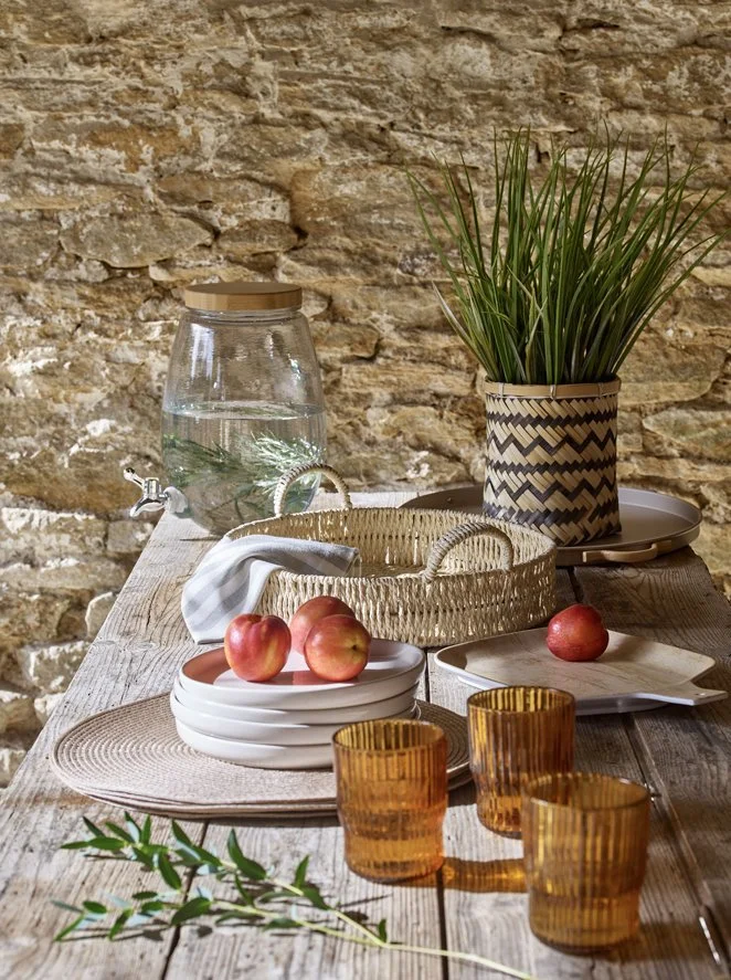 Wooden table with apples on white ceramic plates, amber-colored drinking glasses, a wicker basket, a large glass water pitcher, a potted plant with long green leaves, and a stone wall background.