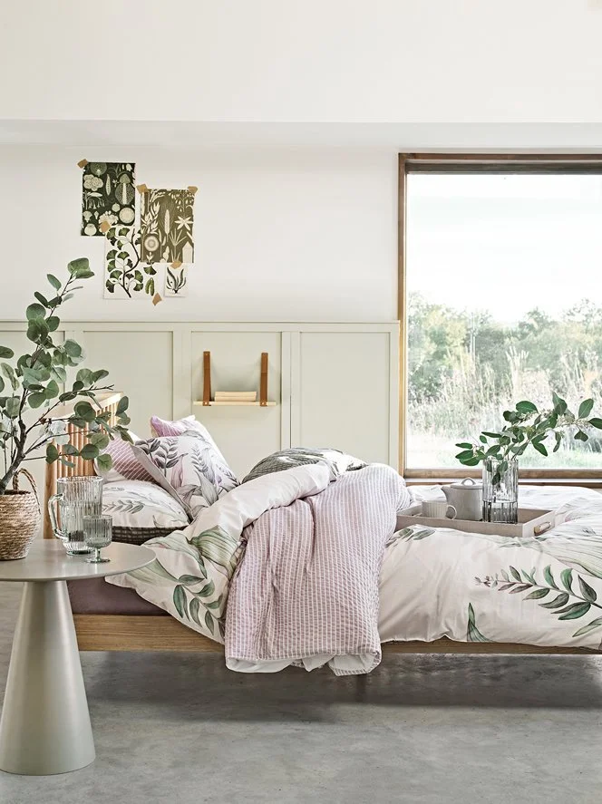 A neatly made bed with floral and pink bedding, a tray with drinks, and green plants in a bright room with a large window and minimal decor.