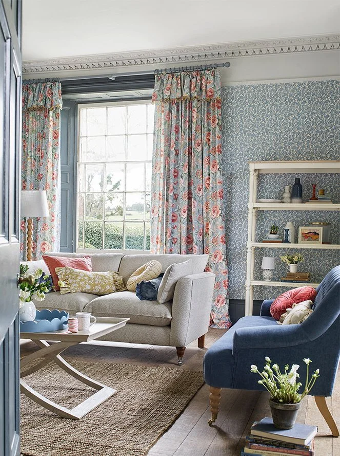 A cozy living room with a large window dressed in floral curtains, a beige sofa with colorful pillows, a blue armchair with a pillow, a small wooden coffee table, a rug, a white bookshelf with decor items, and a potted plant with white flowers.