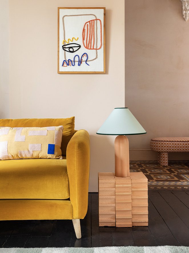 A yellow sofa with a patterned throw pillow, a table lamp with a light green shade and pink base on a wooden block side table, with an abstract art piece hanging on a beige wall above.