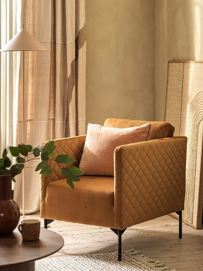 A cozy living room corner with a tan quilted armchair and pink cushion, next to a wooden side table with a potted plant and a small cup, and beige curtains with a light streaming in