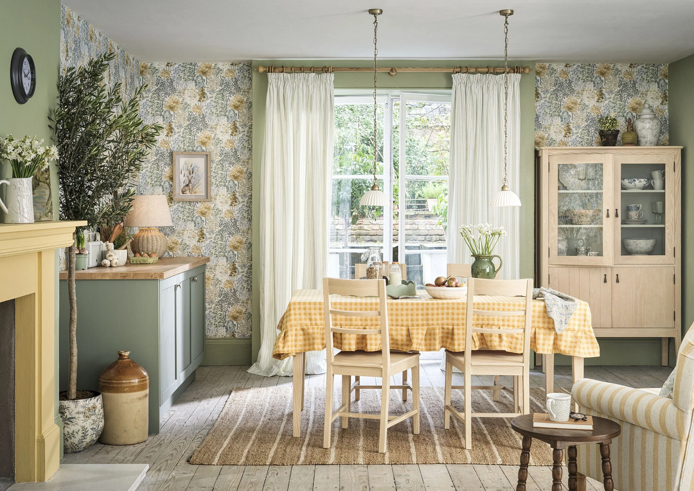 A cozy, well-lit dining room with a wooden table covered with gingham tablecloth, surrounded by wooden chairs. There is a large window with cream curtains, a sideboard with a lamp and decorative items, a glass-front cabinet with dishes, and a small s
