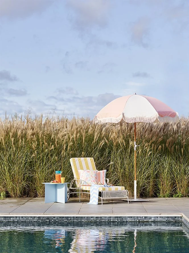 A poolside setup with a striped yellow and white lounge chair, a pink and white umbrella, a small blue table with a drink and snacks, and decorative pillows, surrounded by tall grass and a cloudy sky.