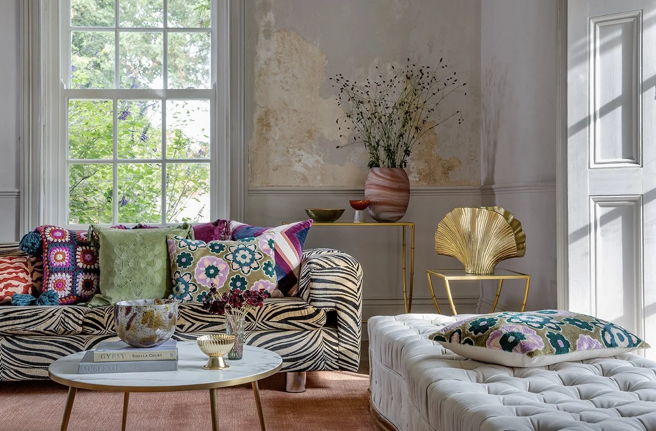 Living room with a large window, decorated with colorful pillows on a patterned sofa, a marble-topped coffee table with books and decorative items, and a white tufted ottoman with a pillow, featuring vintage wall repairs, a pink vase with twigs, and 