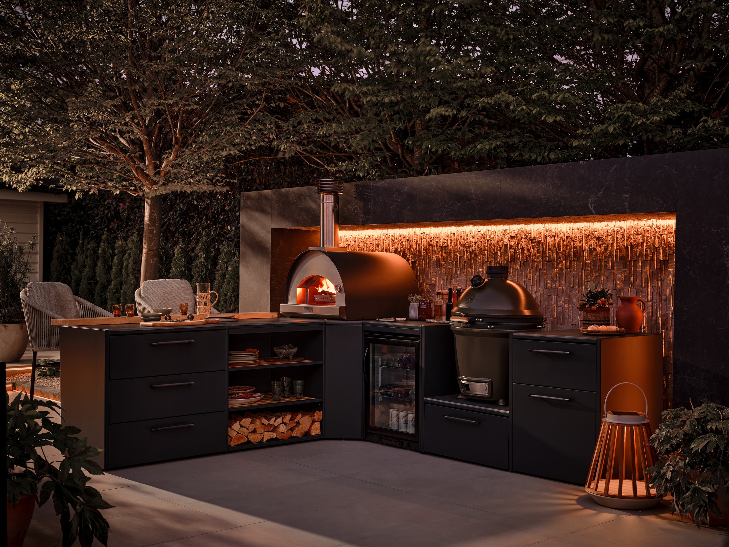 Outdoor kitchen with pizza oven, grill, mini fridge, and dining area against a backdrop of trees and a decorated stone wall illuminated by warm lighting at dusk.