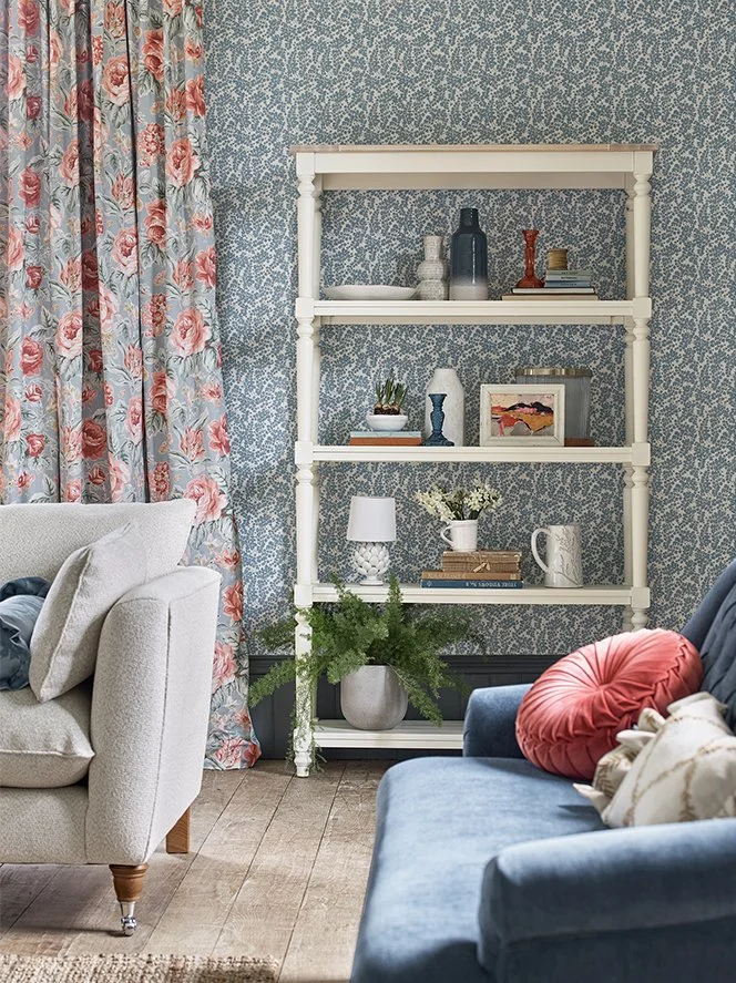 Living room with floral curtains, a cream sofa, a blue sofa with pillows, a white shelving unit with decorative objects, vases, books, a lamp, a picture frame, and a large plant in a gray planter.