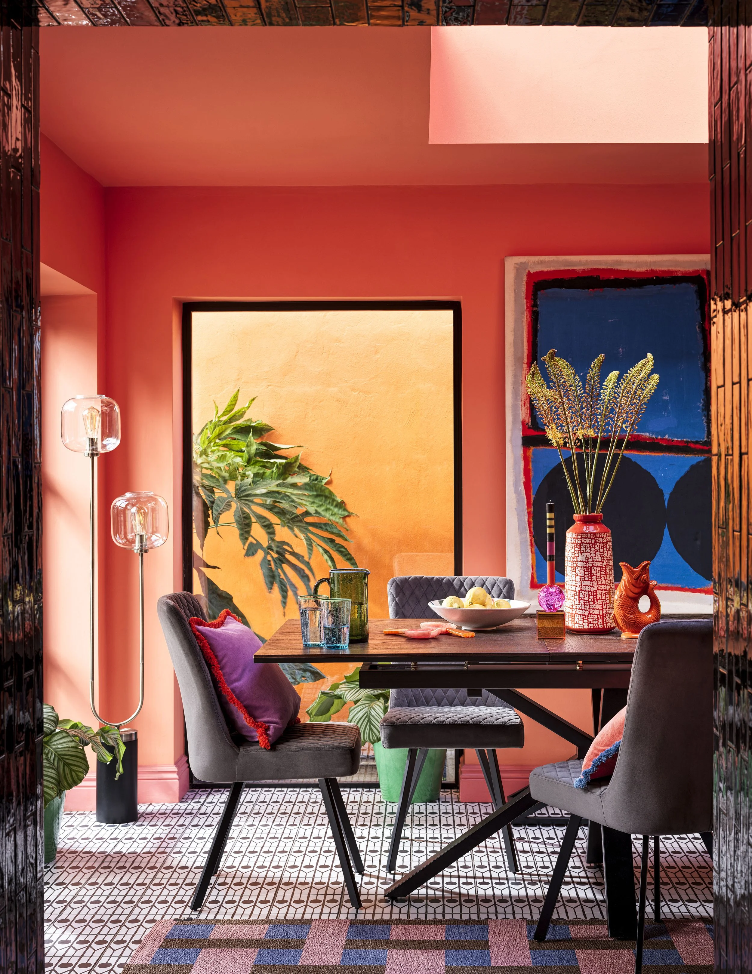 A colorful dining room with orange walls, patterned black and white floor tiles, and a wooden dining table. The table has glassware, a bowl of lemons, and decorative items including a large red and white vase with lavender, a small orange sculpture, 