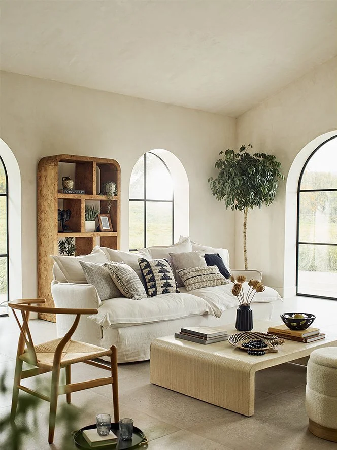 Bright living room with a white sofa covered in neutral-toned pillows, a wooden bookshelf, a potted plant near arched windows, a beige ottoman with decorative items, and a wooden chair with woven seat.