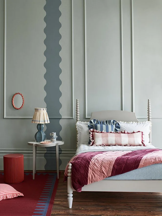 A bedroom with a light gray wall featuring decorative molding and a darker gray vertical stripe; a white bed with pink, white, and blue striped pillows and a red and pink quilt; a gray side table with a blue lamp and a small figurine; a red cylindric