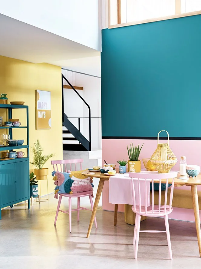 Colorful, modern dining area with pink and green decor, include a pink chair with a plush pillow, a pink tablecloth, potted plants, and decorative woven basket, in a bright, well-lit room.