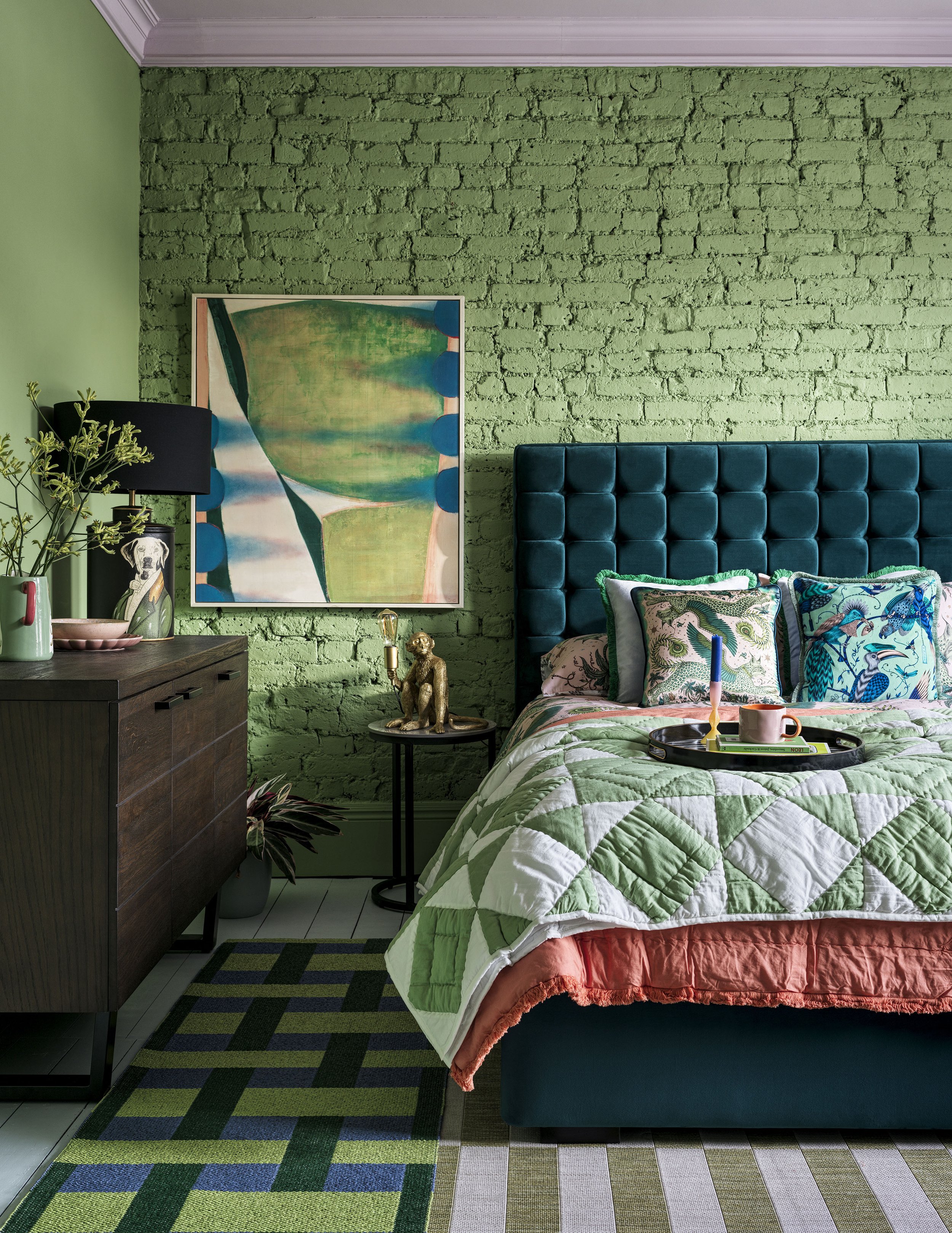 A bedroom featuring a green brick wall, a blue velvet tufted headboard, colorful patterned pillows, a quilt with green and white diamonds, and a tray with a bunch of items on it. A framed abstract painting and artwork are on the wall, along with a si