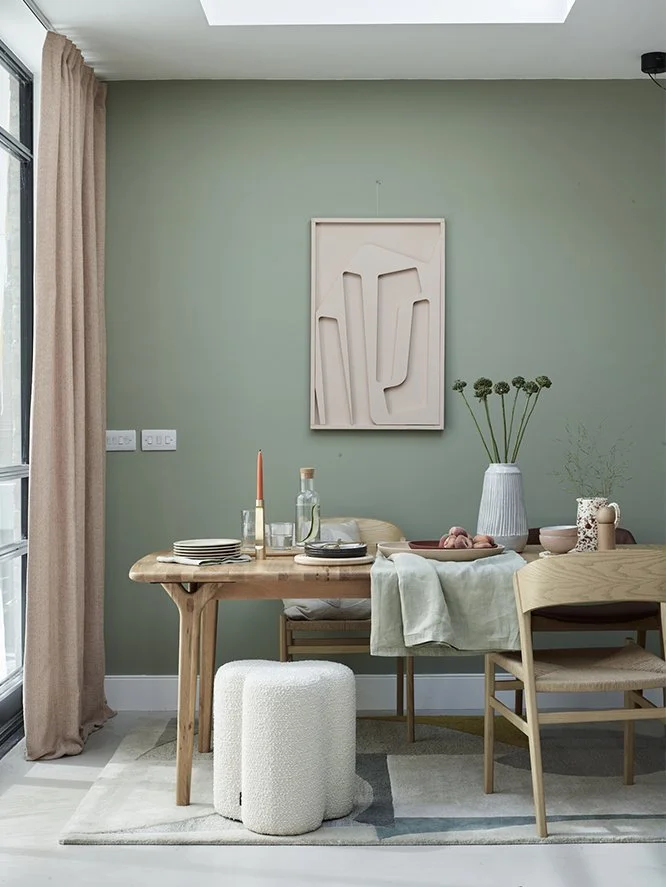 A dining room with a wooden table set for a meal, surrounded by chairs, decorated with a vase of flowers, candles, and tableware. A framed abstract art piece hangs on the green wall. There's a window with beige curtains to the left.