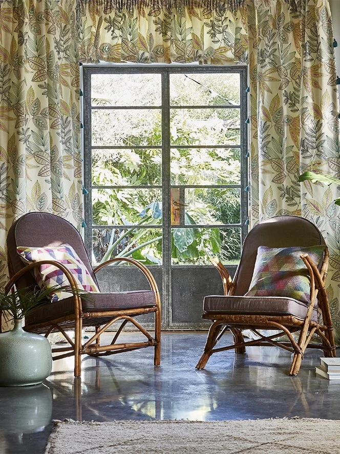 Living room corner with two rattan chairs with cushion pillows, a large window with leaf-patterned curtains, and a potted plant on the floor.
