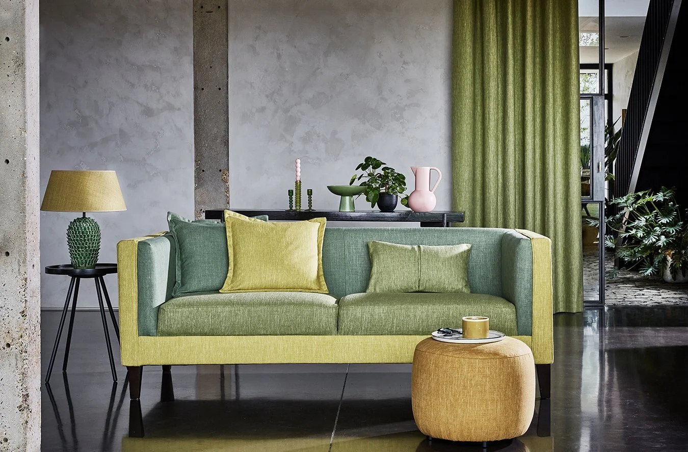 Modern living room with a green and yellow sofa, a small yellow ottoman, a side table with a green cactus-patterned lamp, and a black console table with decorative vases and plants. Green curtains cover a large window.