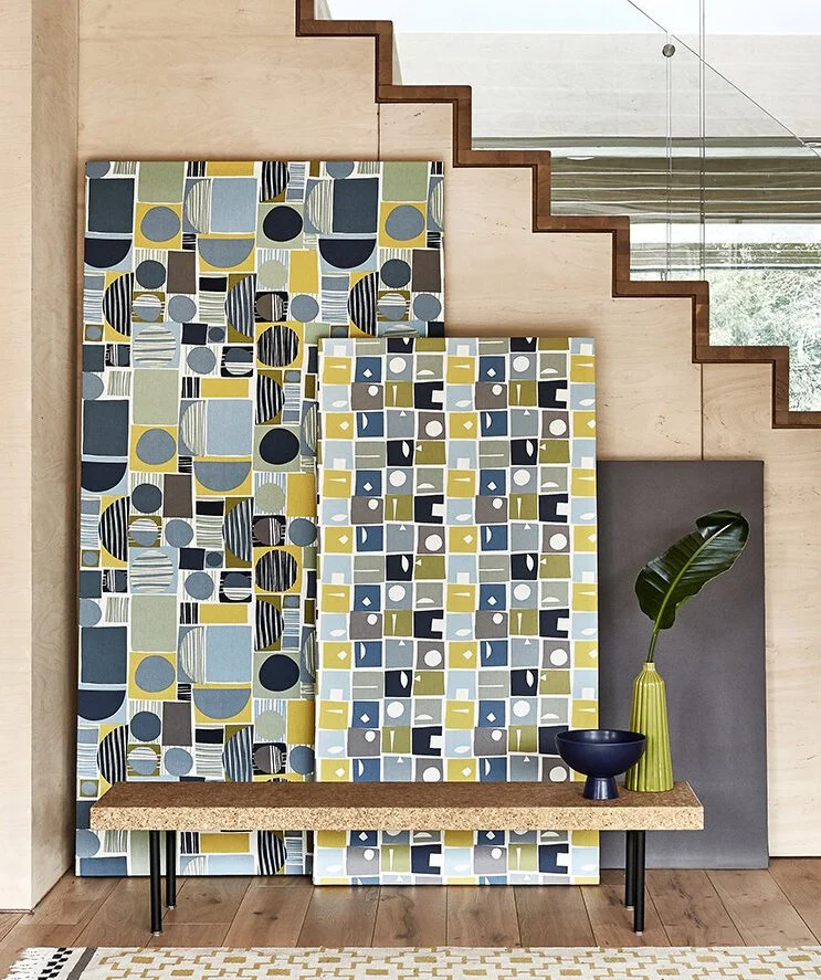 Interior space featuring two large geometric patterned panels with blue, yellow, gray, and black shapes, a wooden bench with black metal legs, and a tall yellow vase with a large green leaf next to a black bowl, against a light-colored wooden wall an