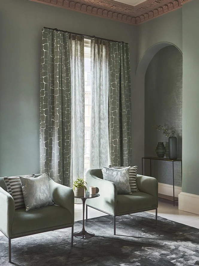 Living room with green armchairs, patterned curtains, a small black side table with a plant and mug, a dark rug, and decorative vases on a console table inside an arched nook.
