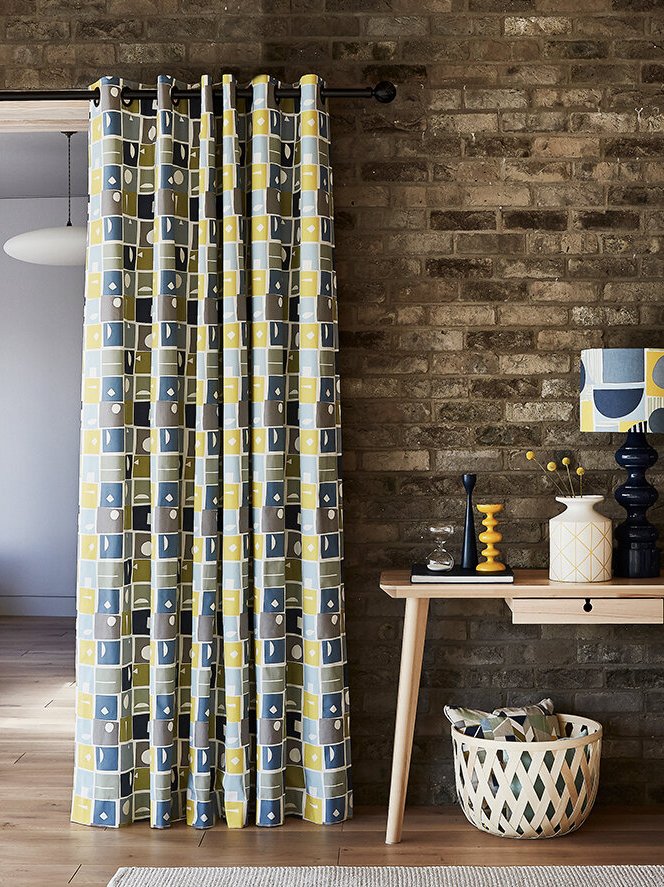 Decorative curtain with geometric pattern in blue, yellow, gray, and white hanging on a black curtain rod in front of a brick wall. A wooden console table with colorful vases, a lamp with a geometric shade, and a woven basket underneath.