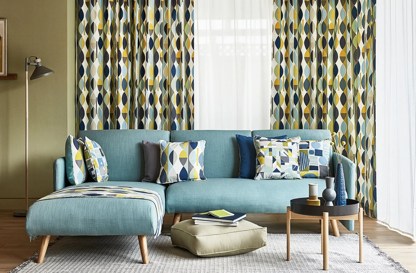 A living room with a turquoise sofa, colorful geometric-patterned pillows, a matching patterned curtain, a beige ottoman with books and a notebook, a round black coffee table with vases and decor, a floor lamp, and a beige rug.