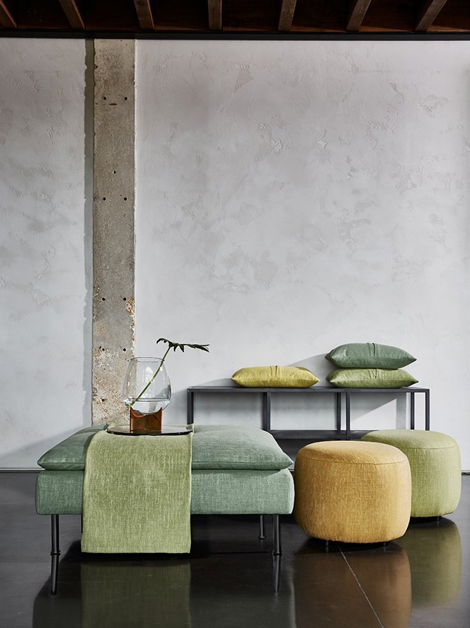 Modern minimalist living room with green and yellow cushions, low black table, and a vase with a single branch, set against a white wall with an exposed concrete pillar.