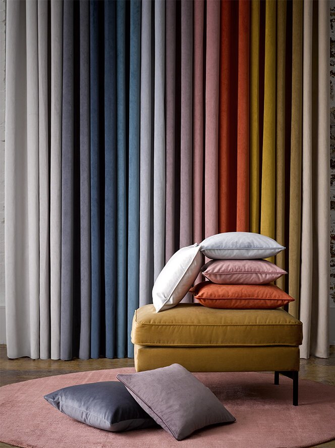 Stacked pillows of various colors on a yellow ottoman in front of multicolored curtains with a pink rug and additional pillows on the floor.