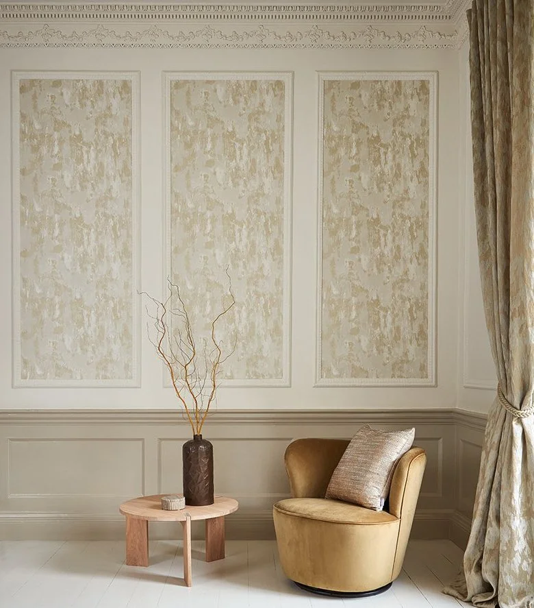 A cozy corner with a beige armchair, a textured cushion, a round wooden side table with a dark vase and dried branches, beige wall panels with textured wallpaper, and a curtain on the right side.
