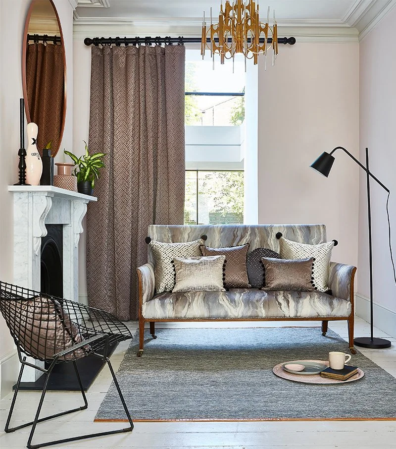Living room with a patterned sofa with multiple pillows, a black floor lamp, a view of trees through the window, a decorative mirror above the fireplace, and a tray with a mug and plate on the floor.